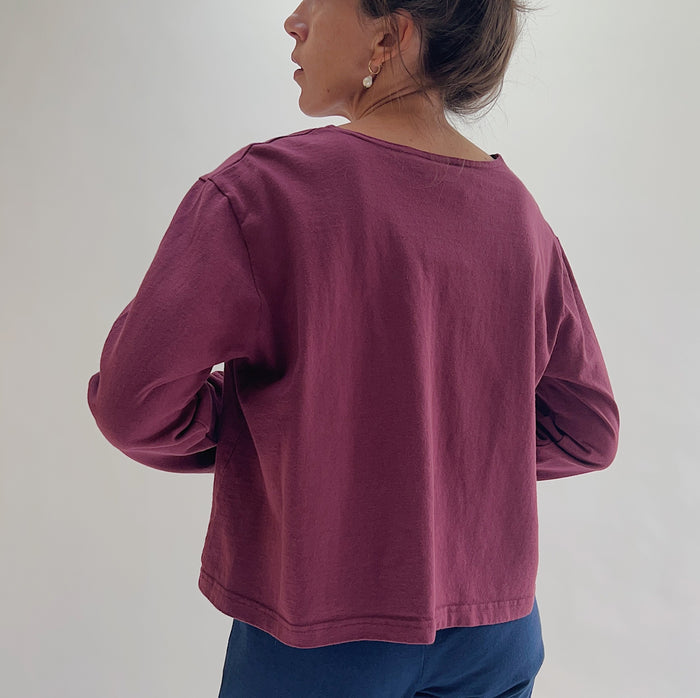 Back view of model wearing Pacific Cotton rhone boxy shirt, showing wide back neckline, long sleeves, boxy cropped silhouette, banded hem, and berry plum color