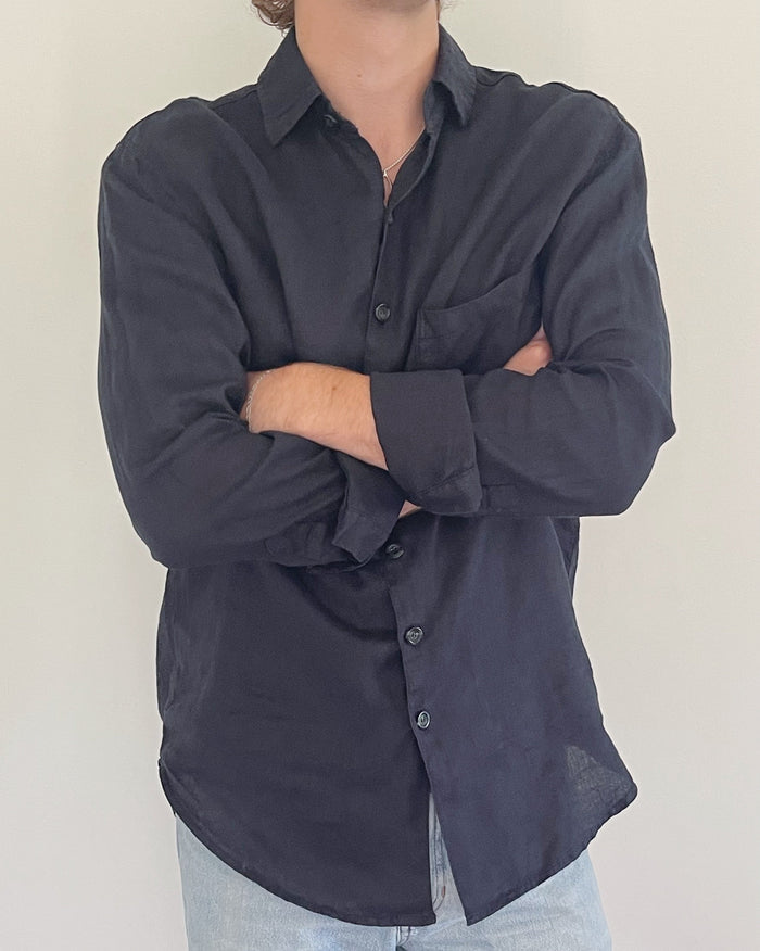 A man wearing a dark navy long-sleeve button-up shirt with a chest pocket, paired with light blue jeans, standing with arms crossed against a plain light background.