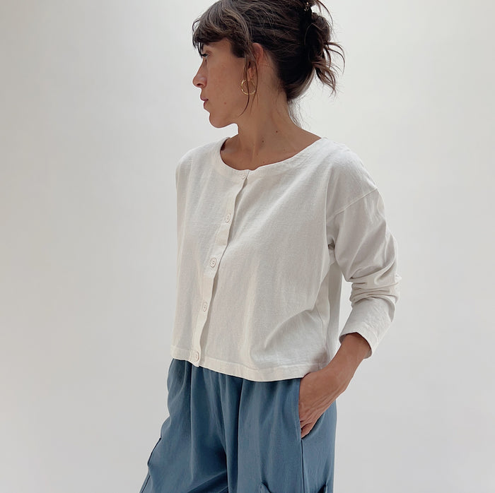 Side three-quarter view of model wearing Pacific Cotton cream button-front shirt, showing crew neckline, button placket, long sleeves, and cropped length layered over blue pants