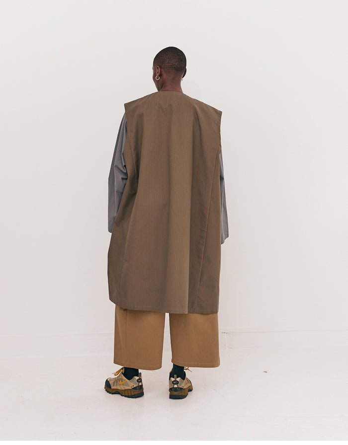 Back view of a model wearing an olive long vest with wide-legged beige pants and brown hiking-style shoes, showcasing minimalist unisex fashion with a focus on volume and clean lines.