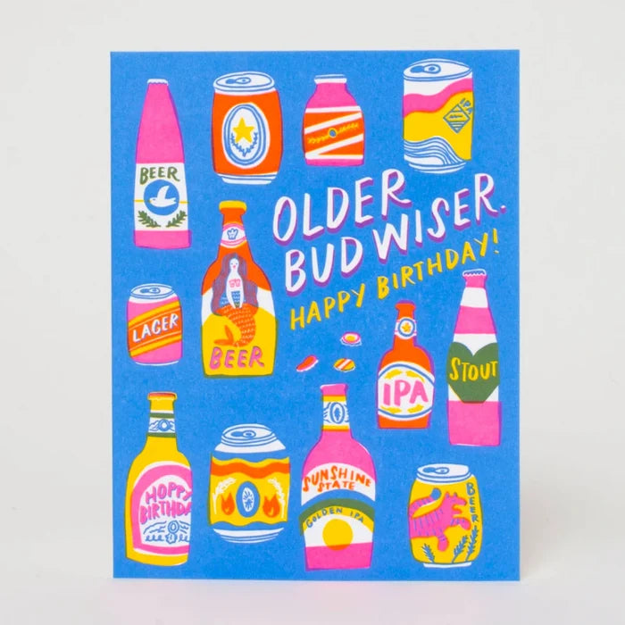 A square blue greeting card featuring colorful illustrated beer bottles and cans around the text 'OLDER BUD WISER. HAPPY BIRTHDAY!' in white and yellow lettering, photographed against a light neutral background.