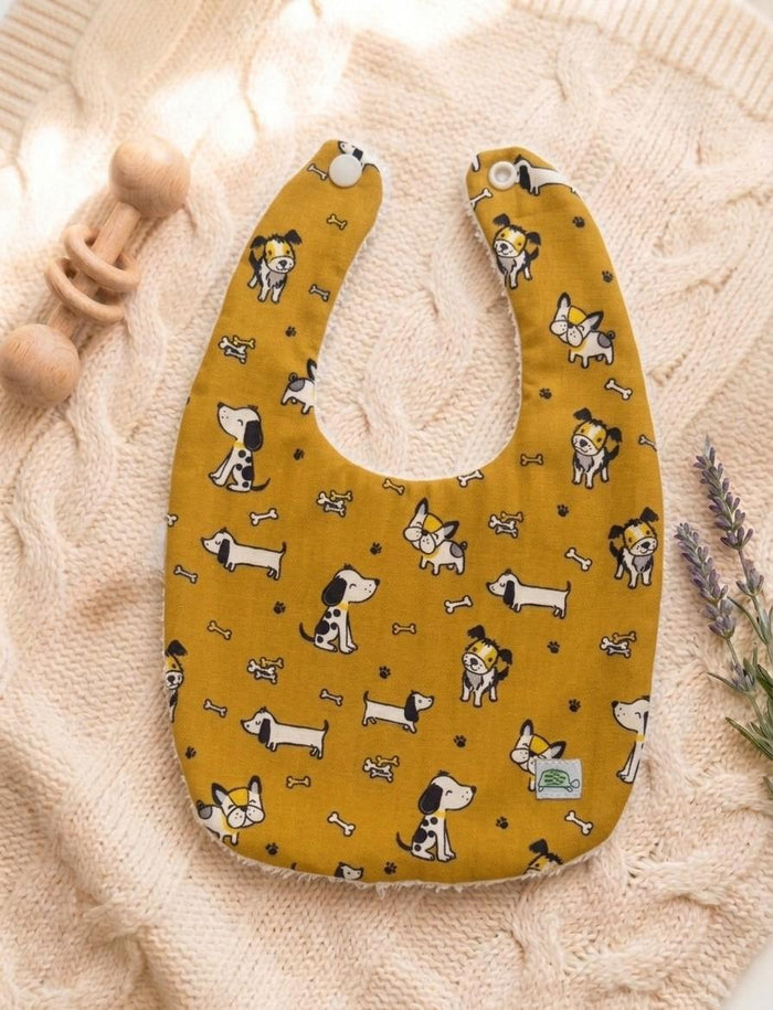 A flat lay of a mustard yellow baby bib featuring a whimsical pattern of various cartoon puppies, tiny bones, and paw prints. The bib has white snap closures at the top and is resting on a soft pink cable-knit sweater alongside a wooden rattle and lavender sprigs.