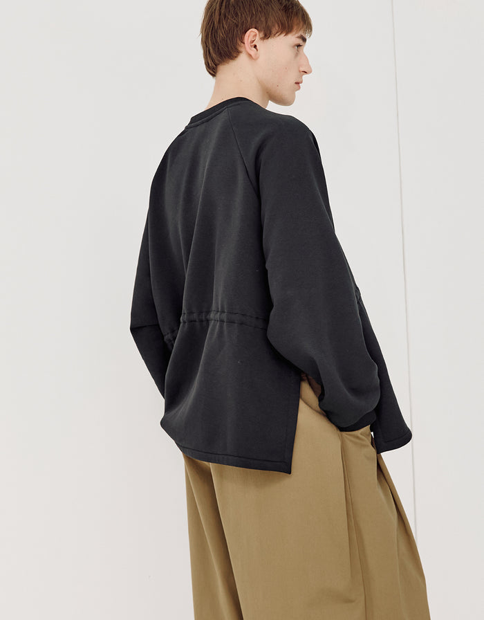 Model wearing a black minimalist sweatshirt with a relaxed fit and subtle asymmetrical hem, paired with loose khaki trousers in a clean, contemporary style.