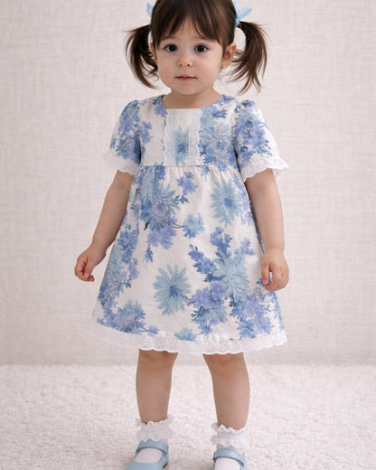 A lifestyle photograph of a young girl with pigtails standing on a soft white rug. She is wearing the Alma dress, a white short-sleeved garment featuring a blue floral print and white eyelet lace trim on the neckline, sleeves, and hem. The look is completed with white ruffle-top socks and light blue Mary Jane shoes against a neutral, textured wall.