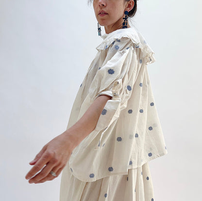A woman wearing a white top with blue floral print with elbow length sleeves a large collar with ruffle trim, viewed from the side.