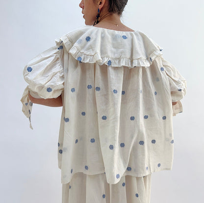 A woman wearing a white top with blue floral print with elbow length sleeves a large collar with ruffle trim, viewed from the back.