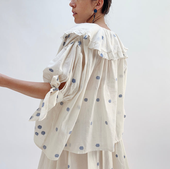 A woman wearing a white top with blue floral print with elbow length sleeves and a large collar with ruffle trim, viewed from the side.