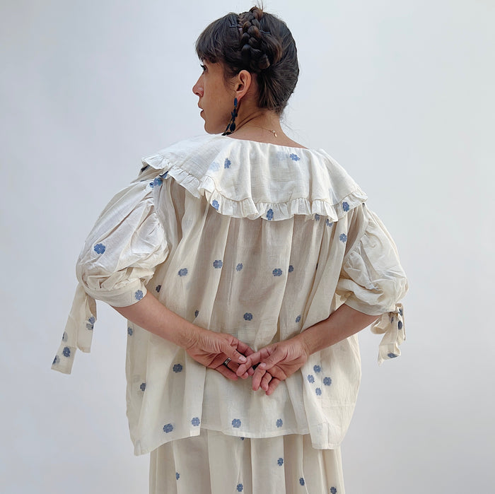 A woman wearing a white top with blue floral print with elbow length sleeves and a large collar with ruffle trim, viewed from the back.
