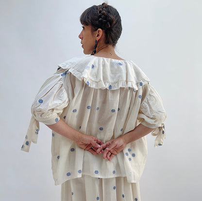 A woman wearing a white top with blue floral print with elbow length sleeves and a large collar with ruffle trim, viewed from the back.