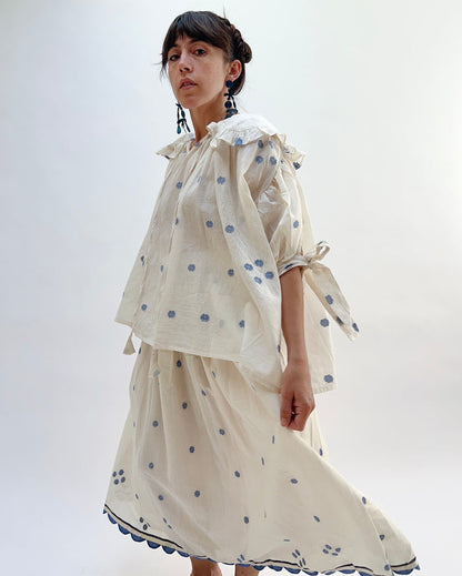 A woman wearing a white top with blue floral print with elbow length sleeves and a large collar with ruffle trim, viewed from the front. She is wearing a matchiing ankle length skirt.