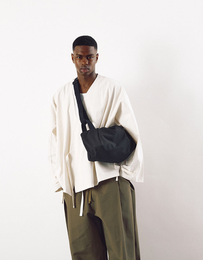 Male model wearing a minimalist cream oversized jacket and olive pants, carrying a black compact crossbody bag with a wide strap, showcasing understated elegance.