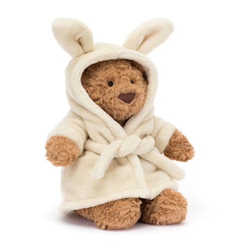 Jellycat Bartholomew Bear wearing a cream plush bathrobe with a bunny-eared hood pulled up, showing golden-brown curly fur face, black bead eyes, and brown nose, seated on a white background