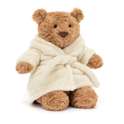 Jellycat Bartholomew Bear wearing a cream plush bathrobe with the hood down, showing golden-brown curly fur, round ears, black bead eyes, and brown nose, seated on a white background