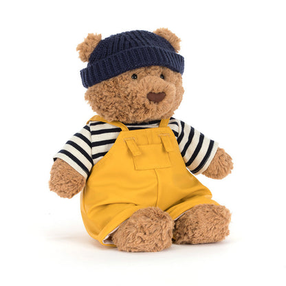 Jellycat Bartholomew Bear wearing a navy ribbed knit beanie, navy-and-cream striped top, and mustard yellow dungarees with buckle detail, seated at a three-quarter angle on a white background
