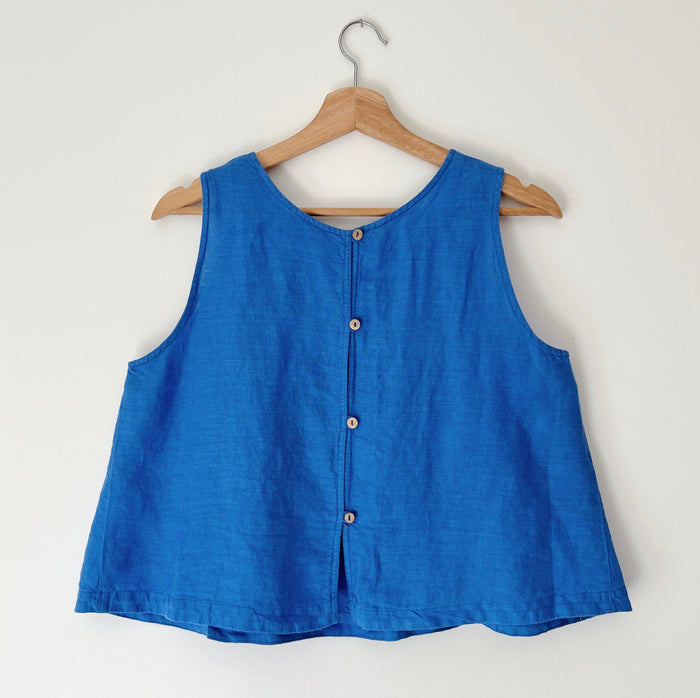 Blue sleeveless tank top on a wooden hanger against a white background. There are wooden buttons down the backside of this tank top.