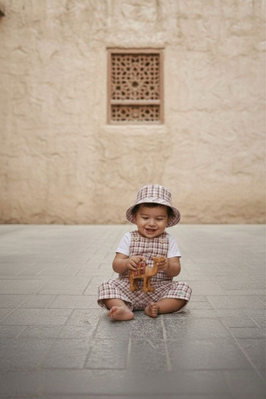 A lifestyle photograph of a smiling baby sitting barefoot and cross-legged on a grey stone-tiled floor. The baby is wearing the Cordoba set, which includes short romper overalls and a matching bucket hat in a red-brown and cream checkered plaid fabric, layered over a simple white short-sleeved t-shirt. The baby is looking down and playing with a small toy camel against a background of a warm, sandy-coloured textured stone wall with an ornate lattice window cutout.