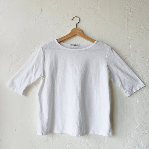 A white elbow sleeve top hanging on a wooden hanger against a textured wall.