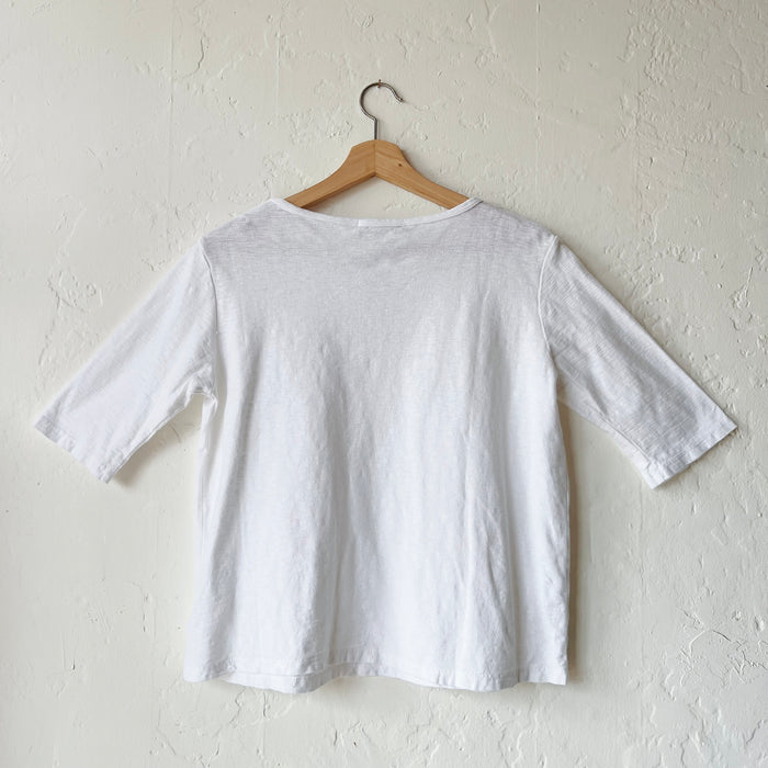 A white elbow sleeve top hanging on a wooden hanger against a textured wall, viewed from the back