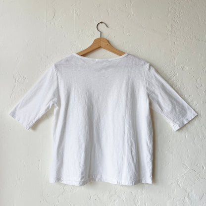 A white elbow sleeve top hanging on a wooden hanger against a textured wall, viewed from the back