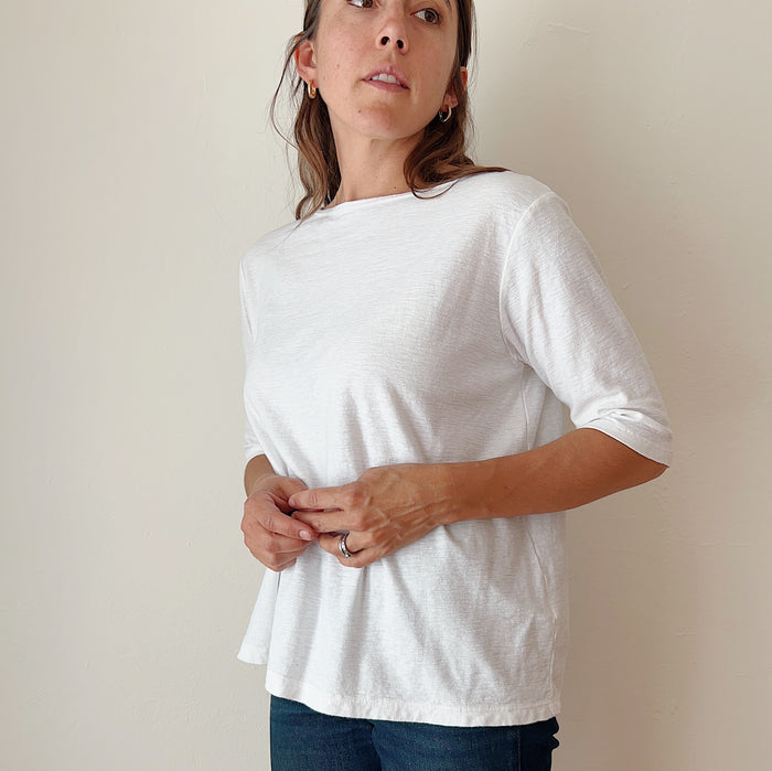 A person standing against a white background, wearing a white elbow sleeve top and blue jeans.