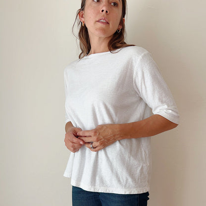 A person standing against a white background, wearing a white elbow sleeve top and blue jeans.