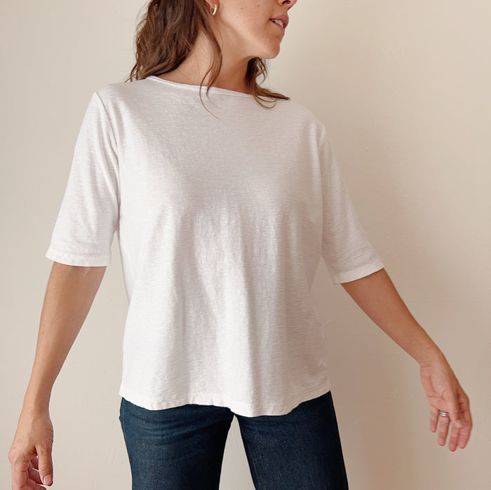 A person standing against a white background, wearing a white elbow sleeve top and blue jeans.