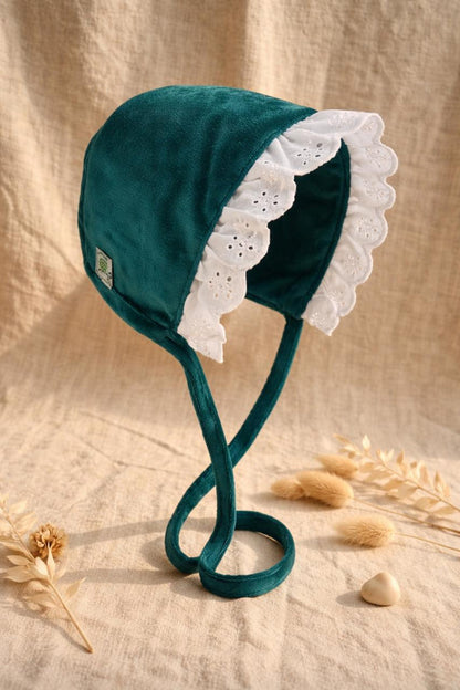 A side-view photograph of a vintage-style baby bonnet made from emerald green velvet. It features a ruffled white eyelet lace trim around the face and long velvet ties, displayed on a neutral linen fabric with dried flowers and a small wooden heart.