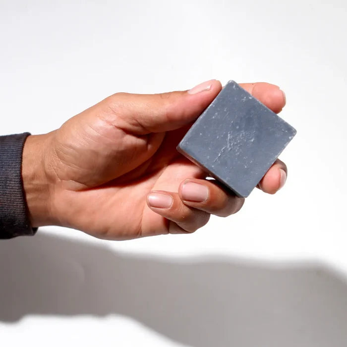 A hand holding a small, square, dark gray bar of soap against a plain white background.