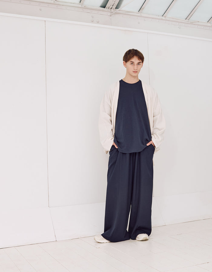 Young person wearing navy wide-leg, wrinkle-resistant pants paired with a loose navy shirt and light beige oversized jacket, standing in a minimalist white space.