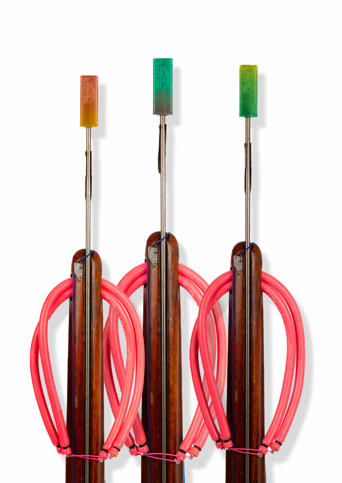 Three Waihana wooden pole spears displayed upright showing flexible tip protectors in orange, teal, and yellow-green colors, each with bright pink rubber bands wrapped around the wooden shafts.