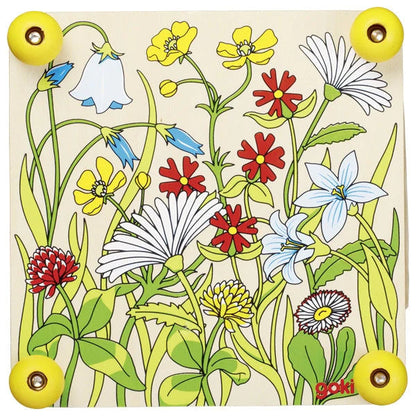 Top view of the Goki Flower Press wooden plate showing a colorful illustrated wildflower meadow with daisies, bluebells, buttercups, clover, and other flowers, with four yellow screw knobs at the corners and the Goki logo in the bottom right