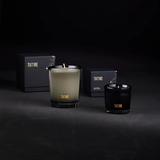 Two Tatine candles, one green and one black, with boxes in the background on a dark surface.