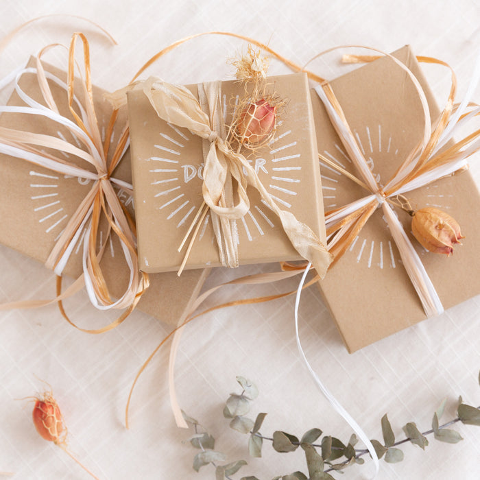 Stack of three gift kraft colored gift boxes with decorative string ties and printed text, displayed on a white surface with eucalyptus leaves and dried flowers scattered around.