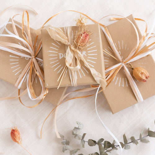 Stack of three gift kraft colored gift boxes with decorative string ties and printed text, displayed on a white surface with eucalyptus leaves and dried flowers scattered around.