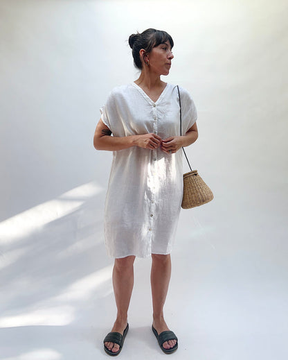 Woman wearing a white dress with buttons down the front and holding a woven bag against a white background