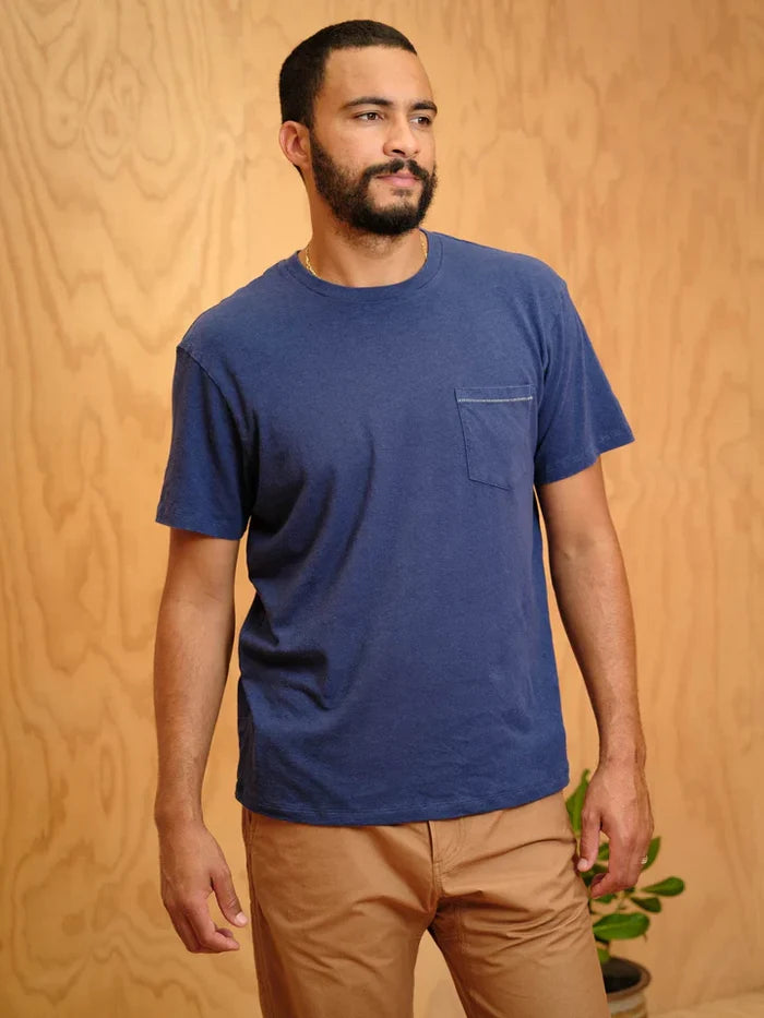Dark haired man with a short beard wearing a blue t-shirt with a pocket against a wooden background