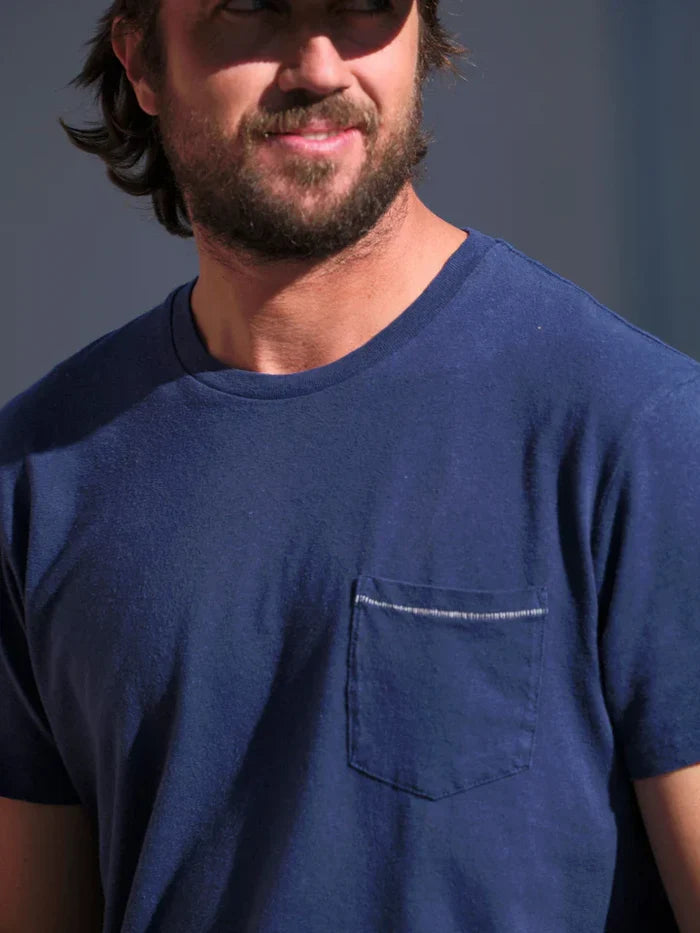 A close-up of a man wearing a navy blue crewneck t-shirt with a chest pocket and white contrast stitching, set against a dark gray background.
