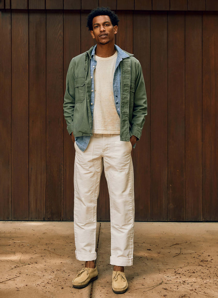 Man wearing the Hencye Canvas Workpant in Natural with a cuffed hem, cream tee, and green field jacket, standing in front of a wood-paneled wall with hands in pockets