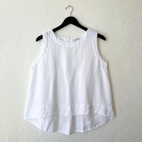White linen high-low tank top by Cut Loose on a hanger, showing the relaxed A-line silhouette, scoop neck, and curved high-low hem