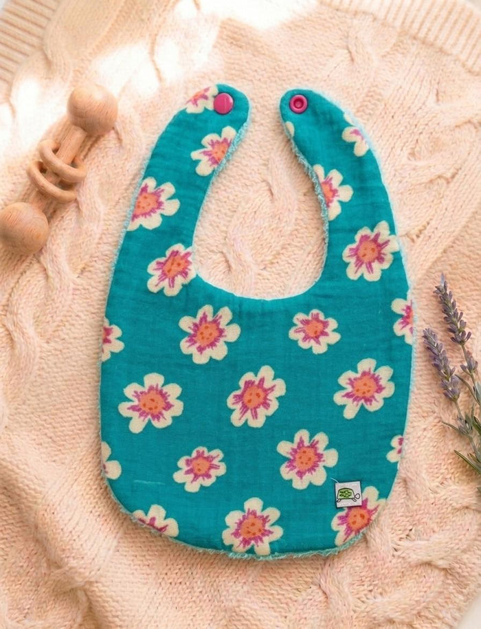 A flat lay of a U-shaped baby bib in a vibrant teal muslin cotton with a white, pink, and orange floral print. The bib features two pink snap closures at the top and a small white turtle logo tag. It is styled on a soft pink cable-knit sweater with a wooden rattle and dried lavender.