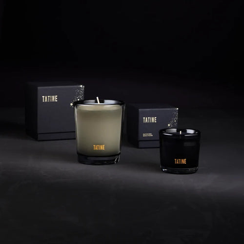 Two Tatine candles, one green and one black, with boxes in the background on a dark surface.