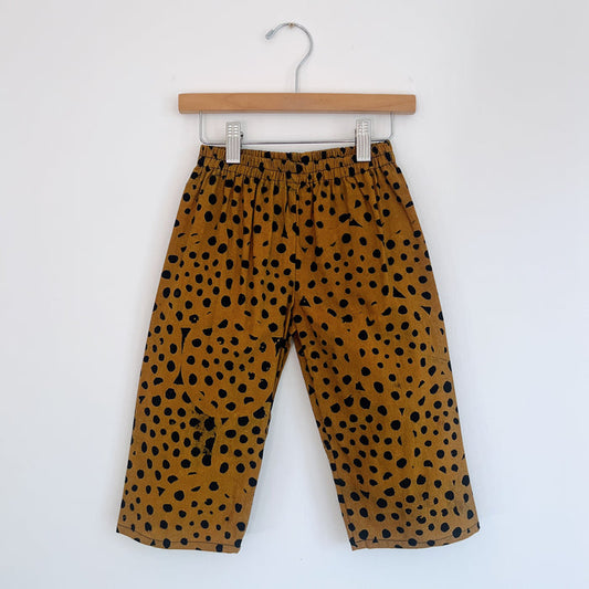Kids lazy pants in cheetah spot Batiki cotton, hanging flat on a wooden clip hanger against a white wall, showing elasticated waistband, golden amber base with hand-printed black cheetah spots, and relaxed fit