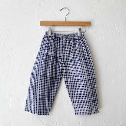 Kids lazy pants in indigo gingham Batiki cotton, hanging flat on a wooden clip hanger against a white wall, showing elasticated waistband and relaxed fit