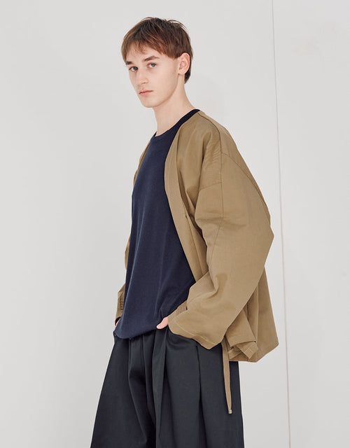 Young man wearing a loose pale brown unisex kimono jacket over a navy shirt and wide black trousers, showcasing minimalist and asymmetric design with relaxed volume.