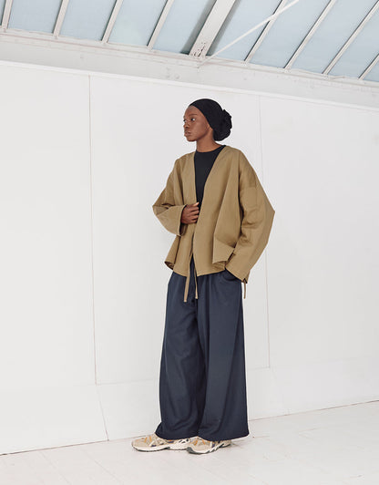 A model wearing a minimalist pale brown oversized jacket with wide sleeves over a black top, paired with loose navy trousers and beige sneakers, in a bright, modern indoor space.