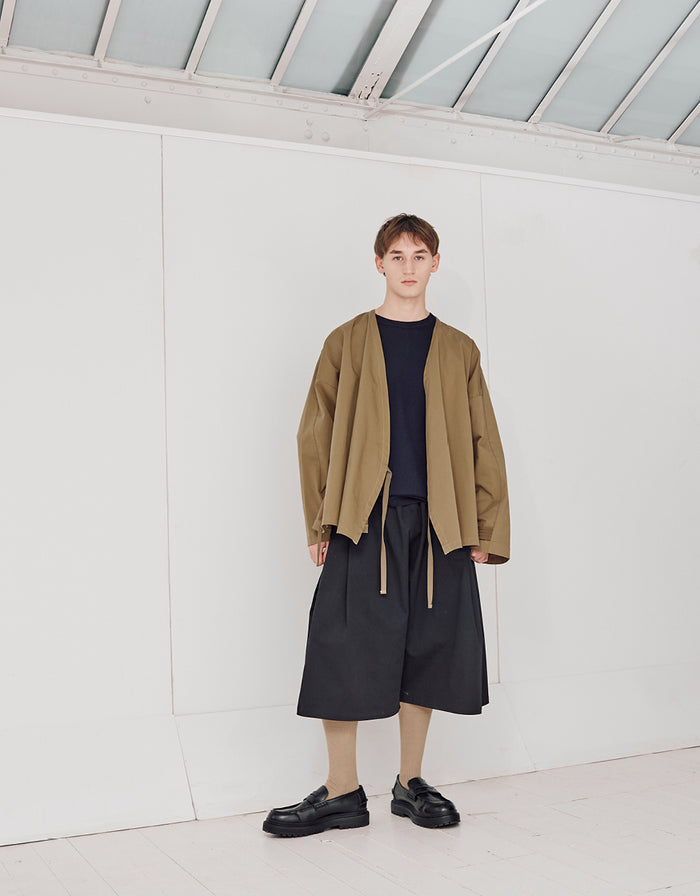 A model wearing a minimalist unisex pale brown kimono-style jacket with wide sleeves, paired with loose black cropped pants and black shoes, standing in a bright, modern space.