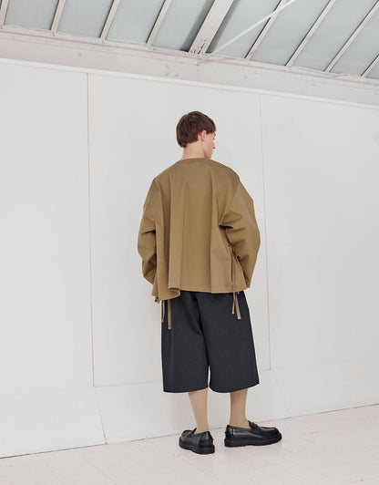 Back view of a model wearing a loose pale brown oversized jacket with wide sleeves paired with black wide-leg cropped pants and black shoes in a minimalist white room.
