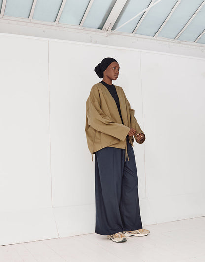 Model wearing a pale brown oversized, minimalist kimono jacket with wide sleeves paired with loose black wide-leg pants and sneakers, standing in a bright modern space.