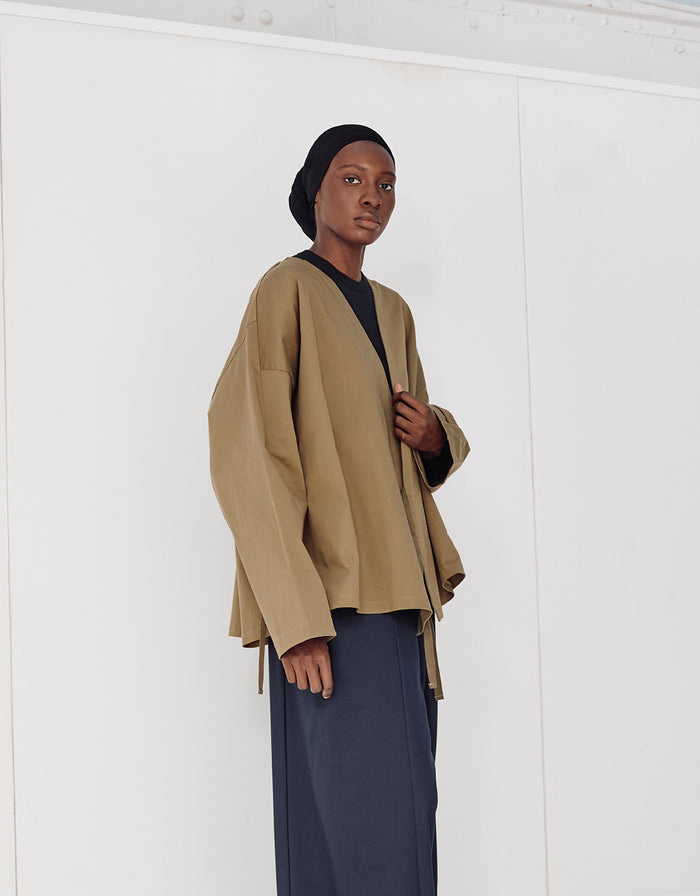 Model wearing a minimalist oversized pale brown kimono-style jacket with wide sleeves, paired with a black top and navy wide-leg trousers against a plain white background.