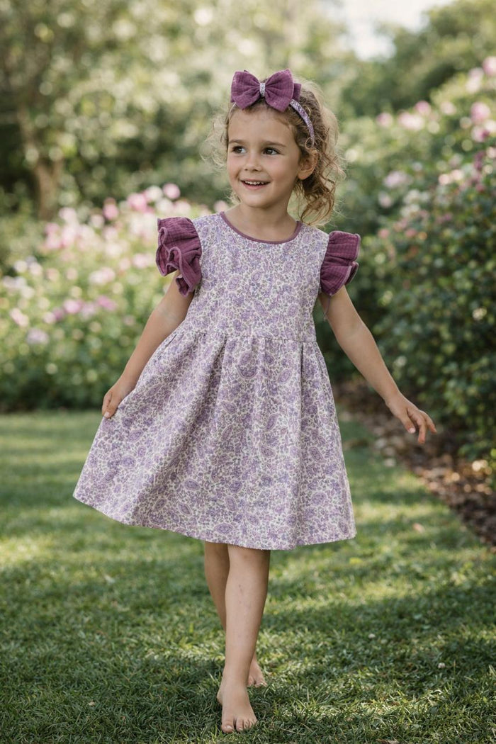 A lifestyle photograph of a young girl with blonde hair walking through a green garden. She is wearing the Larisa dress, which features a light purple paisley print on the body and contrasting deep purple waffle-knit ruffled sleeves. She is also wearing a matching purple waffle bow headband.